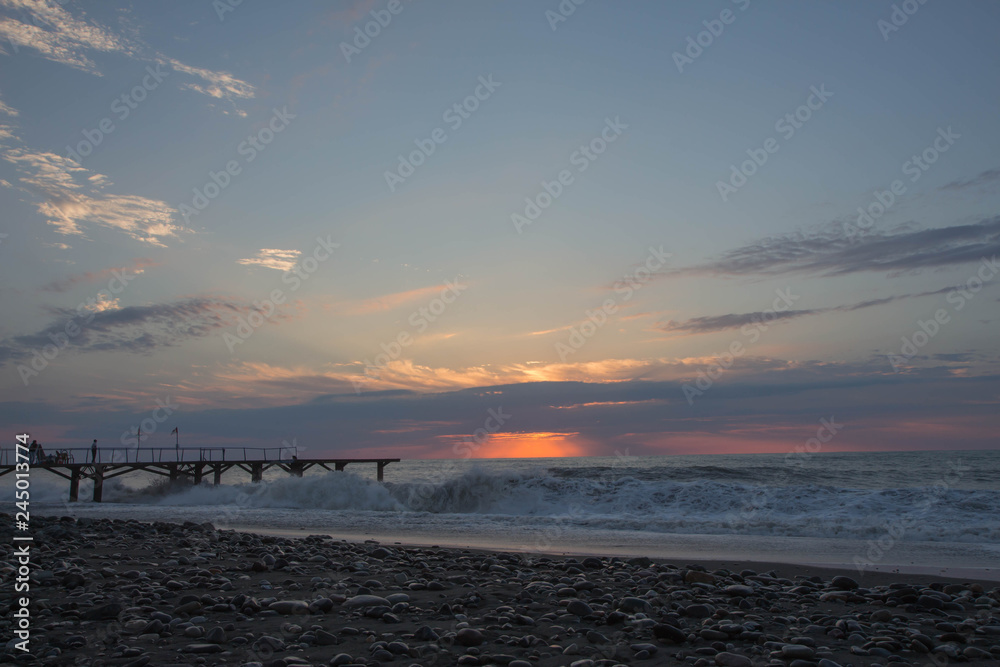 sun setting over Batumi beach pier, as powerful waves roll in, and a very colorful sky is reflected on the beach