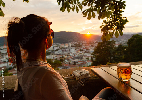 Young woman siting with glass of beer looking at sunset over city Graz in Austria.