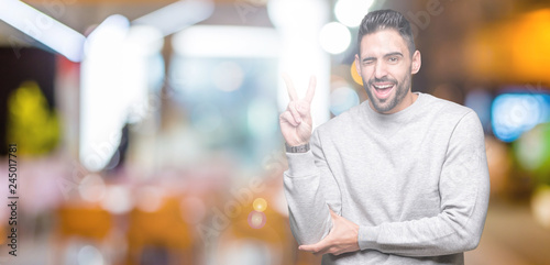 Young handsome man wearing sweatshirt over isolated background smiling with happy face winking at the camera doing victory sign. Number two. © Krakenimages.com