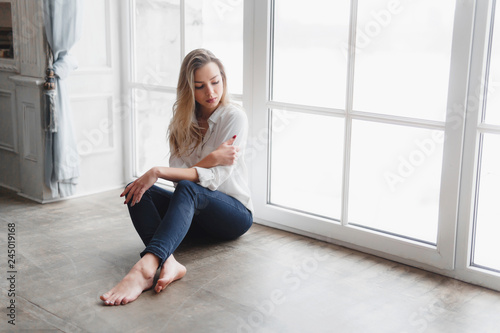 Model blonde white shirt blue pants jeans sitting on wooden parquet floor by window.