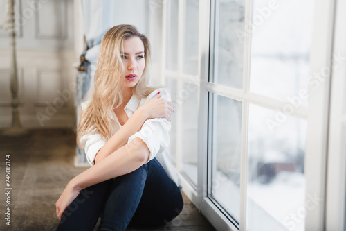 Model blonde white shirt blue pants jeans sitting on wooden parquet floor by window. © NPS Studio