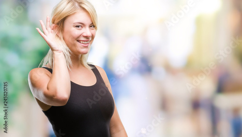 Young beautiful blonde attractive woman wearing elegant dress over isolated background smiling with hand over ear listening an hearing to rumor or gossip. Deafness concept.