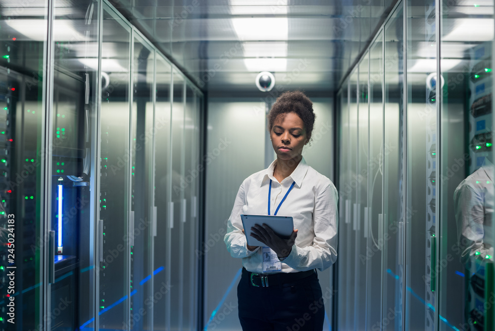 African American woman using tablet while walking in corridor of data center and checking hardware on server racks African American woman using tablet while walking in corridor of data center and checking hardware on server racks