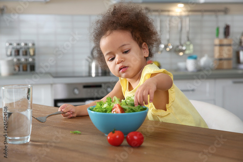 Cute African-American girl eating vegetable salad at table in kitchen