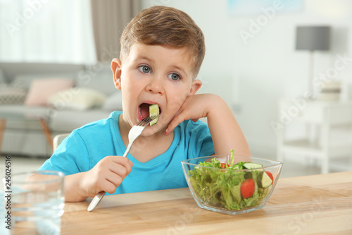 Adorable little boy eating vegetable salad at table in room