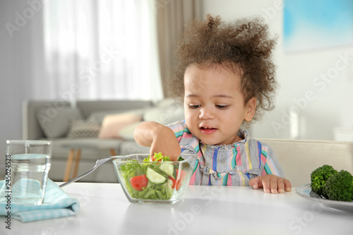 Cute African-American girl eating vegetable salad at table in room
