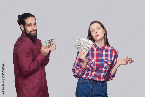 Portrait of rich serious woman with fan of money and near sad poor man, standing and looking at camera. indoor studio shot, isolated on grey background.