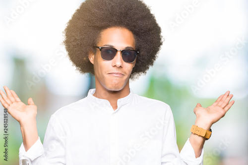 Young african american man with afro hair wearing sunglasses clueless and confused expression with arms and hands raised. Doubt concept.