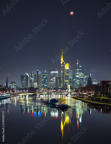 Blood moon during lunar eclipse of over the skyline of Frankfurt  Germany. Reflections on the quiet river Main are being disturbed by a ship.