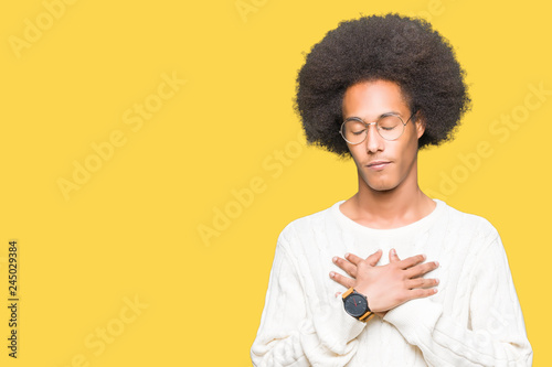 Young african american man with afro hair wearing glasses smiling with hands on chest with closed eyes and grateful gesture on face. Health concept.