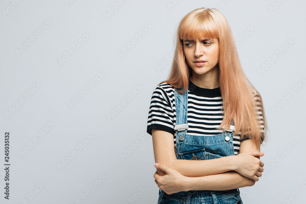 Studio portrait of displeased/angry cute blonde woman in striped t-shirt frowning her face in displeasure and keeping arms folded, looking away. Closed posture, skepticism, disapproving reaction Studio portrait of displeased/angry cute blonde woman in striped t-shirt frowning her face in displeasure and keeping arms folded, looking away. Closed posture, skepticism, disapproving reaction