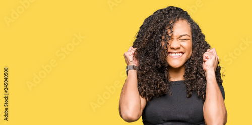 Young beautiful woman with curly hair excited for success with arms raised celebrating victory smiling. Winner concept.