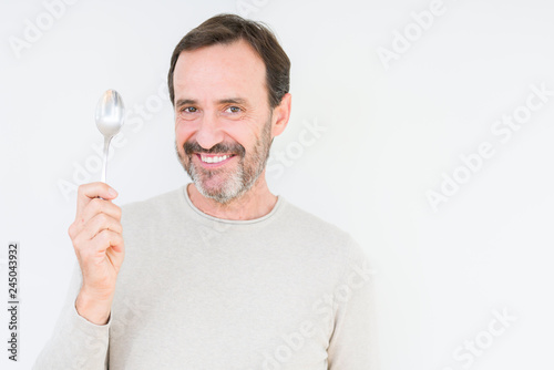Senior man holding silver spoon over isolated background with a happy face standing and smiling with a confident smile showing teeth