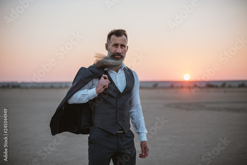 Portrait of bearded mature man in suit walking outdoors on beach during sunset