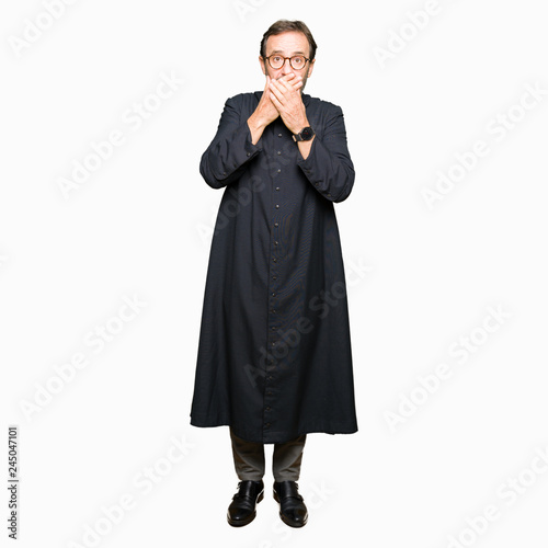 Middle age priest man wearing catholic robe shocked covering mouth with hands for mistake. Secret concept.