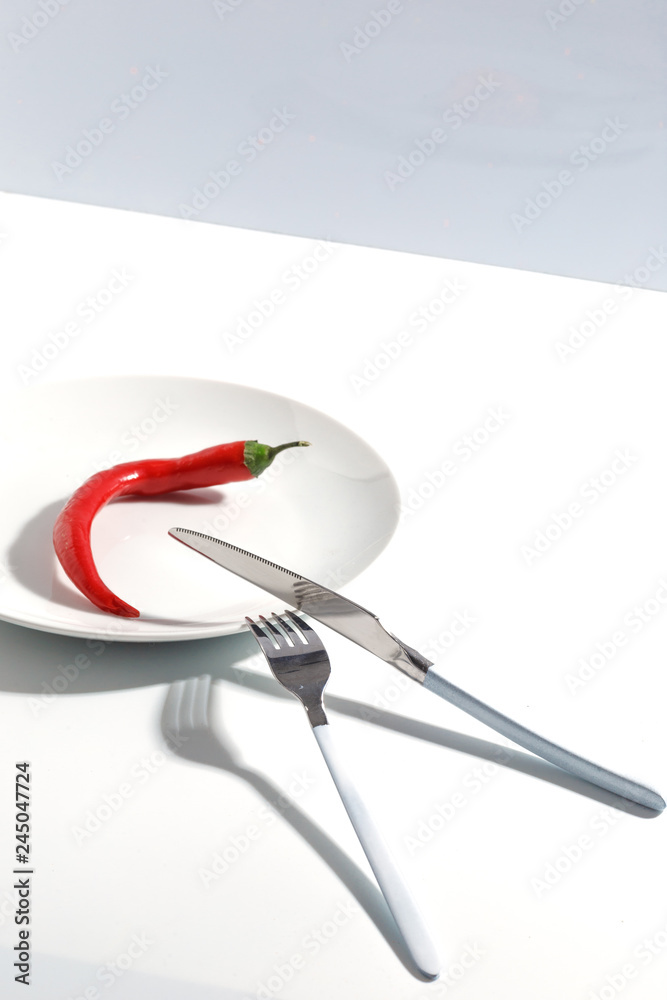 White plate, cutlery and spicy red peppers on a plate on a bright table. Cretive minimalistic concept. White plate, cutlery and spicy red peppers on a plate on a bright table. Cretive minimalistic concept.