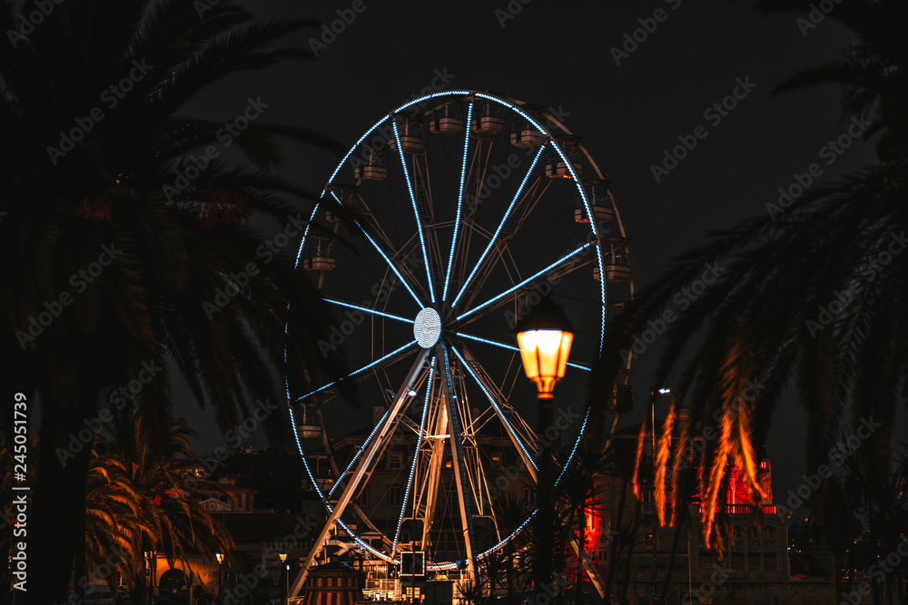 Night shot with a beautifully illuminated Ferris wheel framed with dark silhouettes of palms and a single street lantern in the foreground, Cascais, Portugal