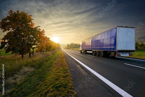 Blue truck driving on the asphalt road towards sunset around line of trees in misty autumnal landscape
