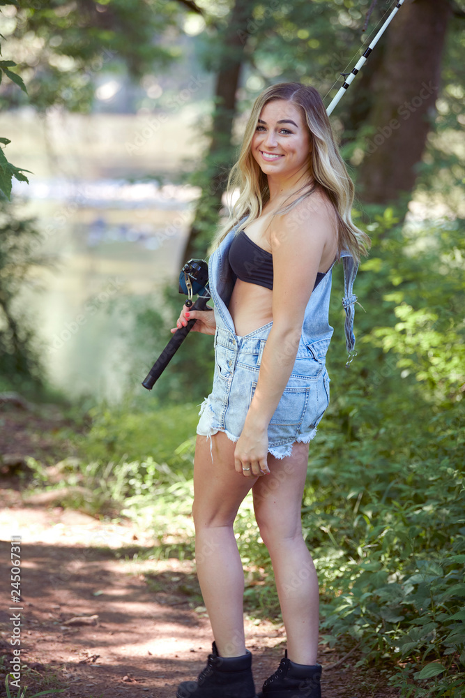 Beautiful blonde young woman fishing near creek wearing coveralls - walking  down trail with rod over shoulder Stock Photo