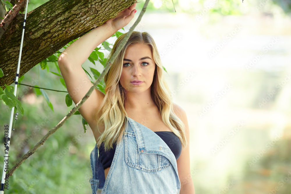 Beautiful blonde young woman fishing near creek wearing coveralls -  standing next to tree Stock Photo