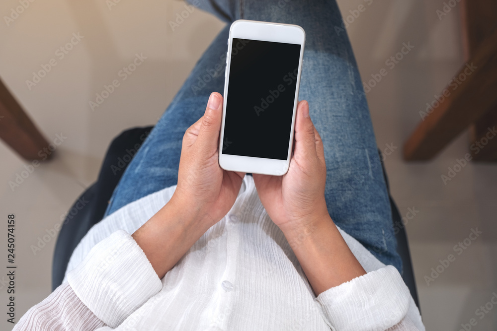 Top view mockup image of hands holding white mobile phone with blank black screen while sitting