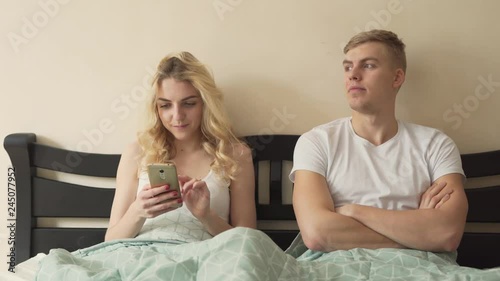 The married couple chills in the bed. The wife plays on her phone and does not pay attention to her husband. He glances into the window, opposite direction and at her phone screen, as he is bored. photo