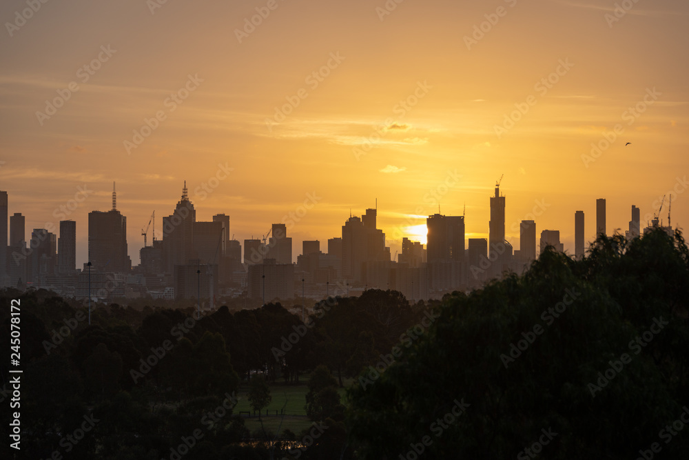 Melbourne city skyline with a glowing orange sky and park trees in the foreground
