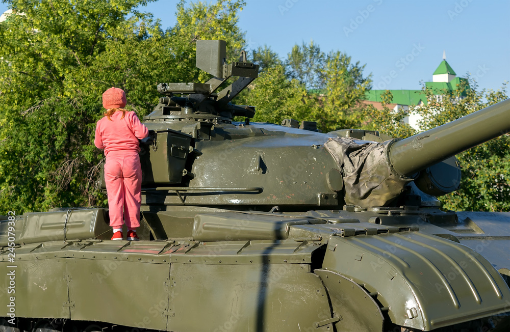 A child, a little girl, examines a military tank in the Park in the urban area A child, a little girl, examines a military tank in the Park in the urban area
