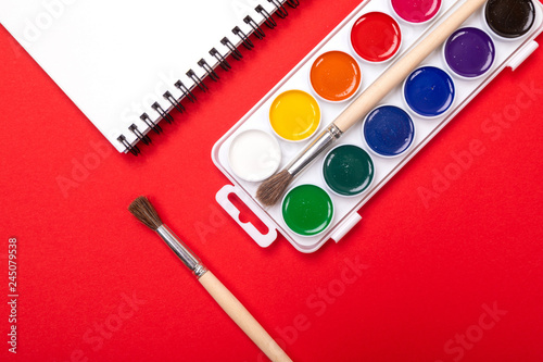 Watercolor paints and brushes with drawing album isolated on red background top view
