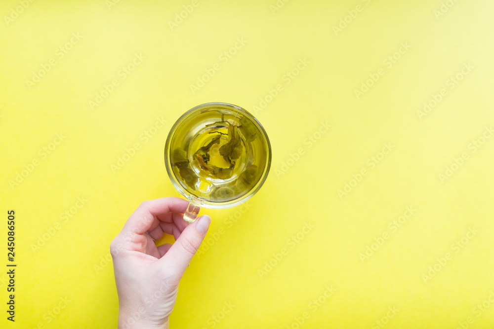 woman's hand holding cup of green tea - top view of minimal flat lay on yellow background.