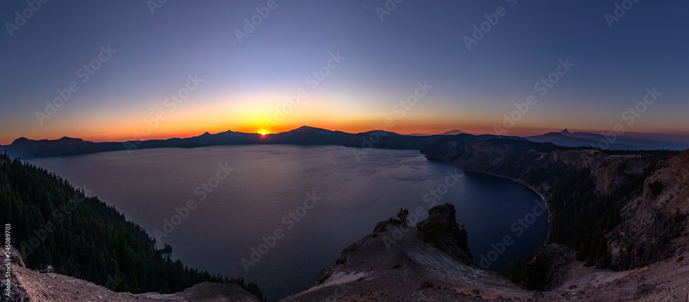 Wide angle panorama of Crater Lake at sunset. Setting sun cast light of red, yellow and orange around sharp rocks of a caldera and surrounding hills