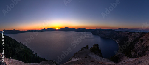 Wide angle panorama of Crater Lake at sunset. Setting sun cast light of red  yellow and orange around sharp rocks of a caldera and surrounding hills