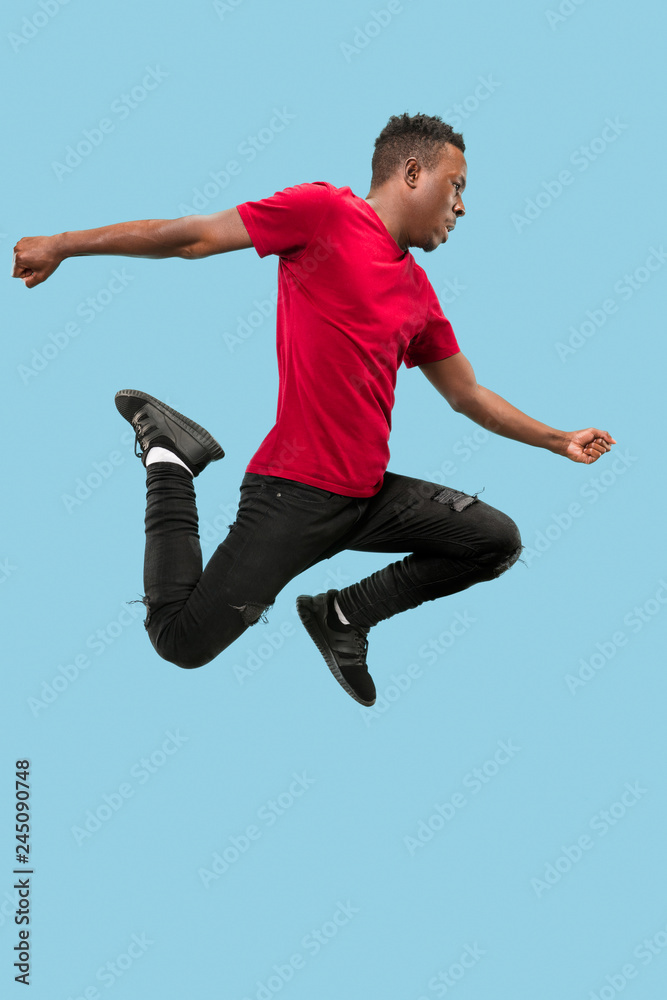 Fototapeta premium Freedom in moving and forward motion. The happy surprised young african man jumping a against blue studio background. Runnin man in motion or movement. Human emotions and facial expressions