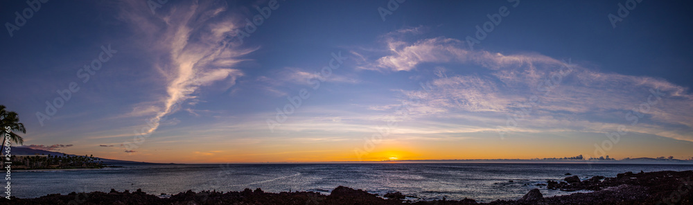 Scenic stitched panorama of a sunset with a dramatic sky over Pacific ocean, Big Island, Hawaii. Orange, yellow and red color over the horizon with a beautiful clouds in a blue sky.