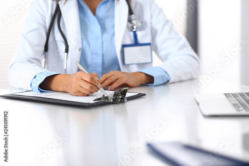 Unknown doctor woman filling up medical form while sitting at the desk in hospital office. Physician at work. Medicine and health care concept