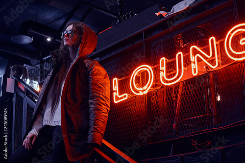 Young stylish girl wearing sunglasses with a coat covering his shoulders standing on stairs at underground nightclub with industrial interior