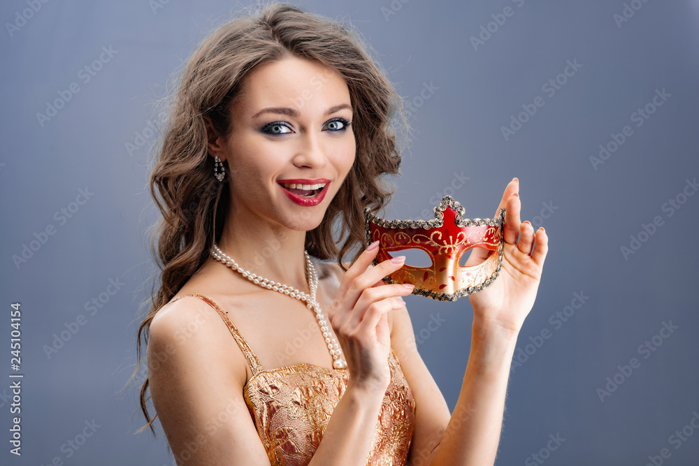 Young woman in a pearl necklace surprised looking at the camera and showing a carnival mask Young woman in a pearl necklace surprised looking at the camera and showing a carnival mask