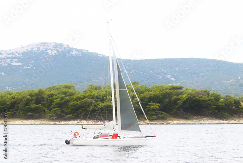 A modern cruise sailing yacht with a Bermuda sloop-type rig goes past the green coast of the Croatian Riviera on a sunny summer day. Adriatic Sea of the Mediterranean region. District of Dalmatia