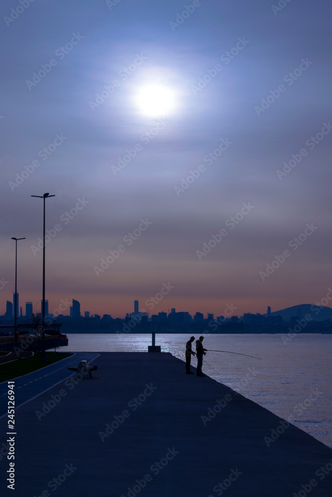 Silhouettes of two fishermen with fishing rods on cloudy morning. Sea and city view on the background. Istanbul. Vertical image