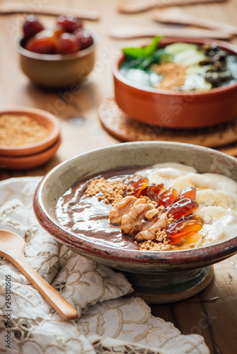 Cocoa Acia bowl with slices banana, fresh dates, candied walnuts and bee pollen topping on a brown wooden background with embroided napkins and wooden spoons photo