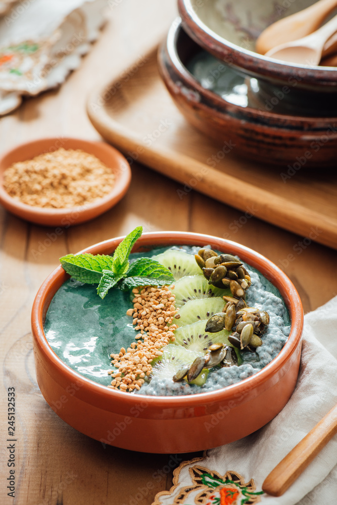 Fototapeta premium Spirulina Acia bowl with toppings of candied pumpkin seeds, kiwi fruit, bee pollen and fresh mint, on a brown wooden background