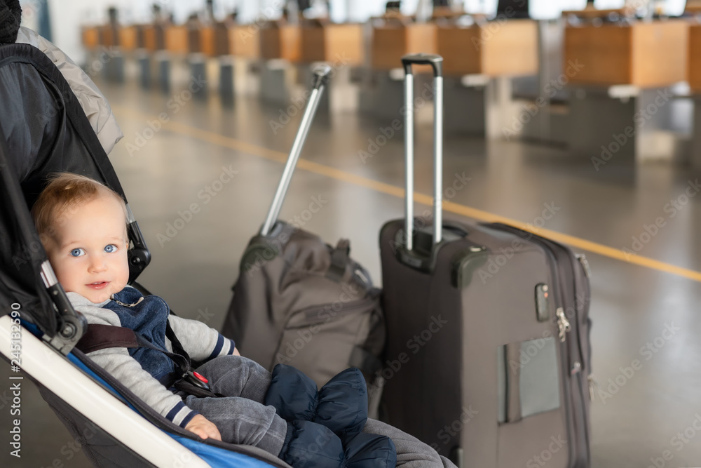 Cute funny caucasian baby boy sitting in stroller near luggage at airport terminal. Child sin carriage with suitcasese near check in desk counter. Travelling with small children concept Stock Foto Ado...