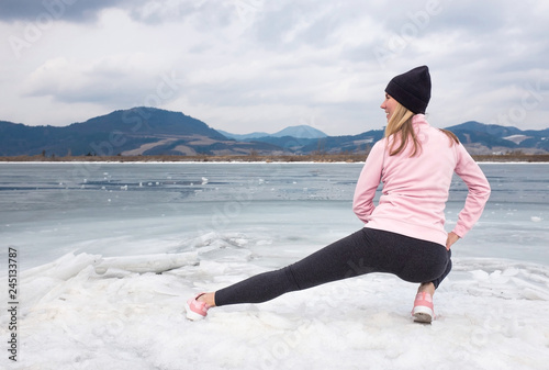 Woman after run does warming up on bank of frozen lake on background of winter mountains photo