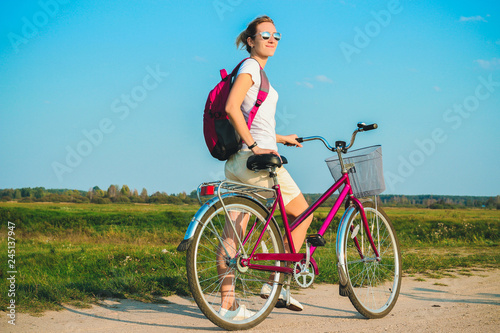 Young beautiful woman standing with her pink bicycle closeup in the summertime on the green fields background