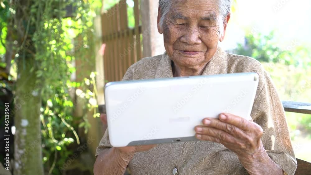 Happy Asian old woman / grandmother smiling and using tablet pointing finger on touching screen for typing chating sms message to Grandchildren