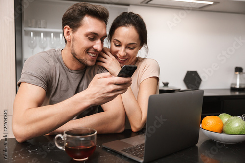 Young couple man and woman using laptop with smartphone, while sitting in kitchen