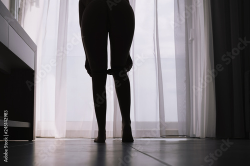 Bottom view of silhouette of beautiful woman with sexy legs taking off her stockings in a room against the backdrop of curtains © flowertiare