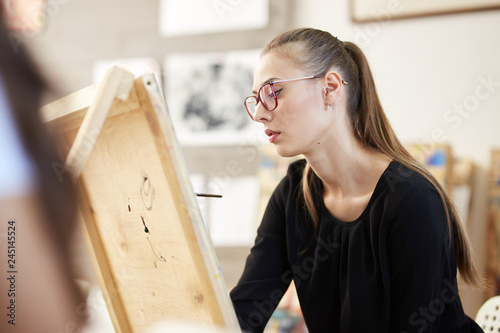 Pretty fair-haired girl in glasses dressed in black blouse sits at the easel and paints a picture in the art studio