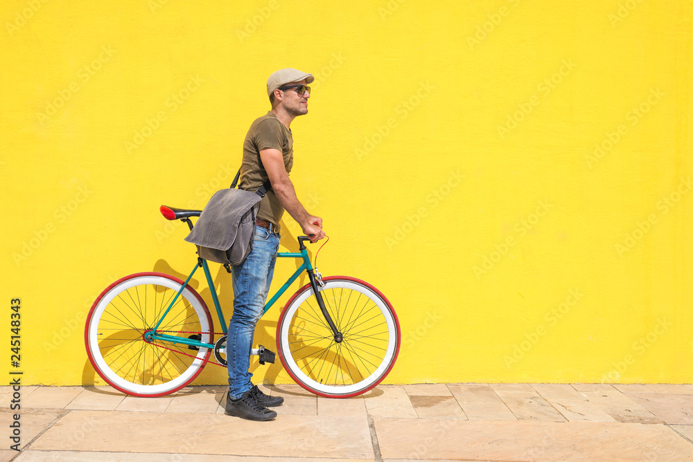 Fototapeta premium Side view of a young hipster man with a fixed bike wearing casual clothes while looking away against a yellow wall outdoors in a sunny day