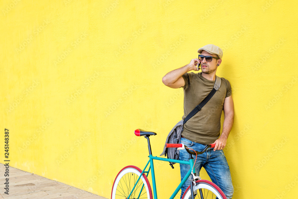 Naklejka premium Side view of a young adult man with a vintage bike and wearing casual clothes and sunglasses standing against a yellow wall while using a mobile phone in a sunny day